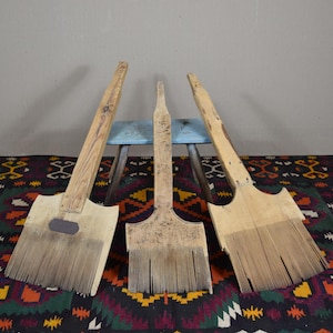May include: Three vintage, large paint brushes with wooden handles and bristles. The brushes are arranged against a gray wall and a colorful patterned rug. The brushes are of varying sizes and show signs of wear.