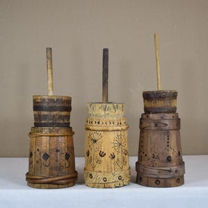 Könnte beinhalten: Drei antike Butterfässer aus Holz mit zylindrischen Körpern und langen Holzstempeln. Die Fässer sind in verschiedenen Braun- und Beigetönen gehalten und weisen dekorative Schnitzereien und Metallbänder auf. Sie werden auf einer weißen Oberfläche präsentiert.