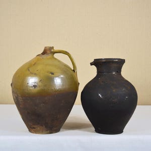 Puede incluir: Dos jarras antiguas de cerámica. Una es una jarra grande y bulbosa con asa, esmaltada en tonos verde oliva y marrón. La otra es una jarra más pequeña, de color marrón oscuro, con cuello estrecho y cuerpo redondeado. Ambas se exhiben sobre una superficie blanca.