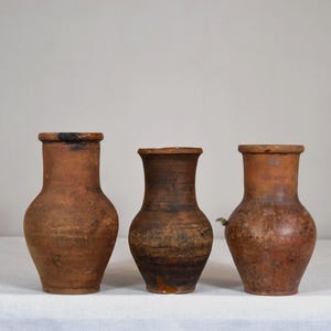 Puede incluir: Tres jarrones antiguos de terracota con un aspecto rústico y envejecido. Los jarrones varían ligeramente en forma y tamaño, con una paleta de colores cálidos y terrosos. Los recipientes tienen una forma clásica y redondeada, perfectos para flores secas.