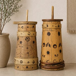 Op de afbeelding: Twee antieke houten botervaten. De vaten zijn cilindrisch met een uitlopende basis en een deksel met een houten handvat. Ze zijn versierd met donkere, gestileerde bloemen en geometrische patronen. Het hout heeft een warme, natuurlijke tint.