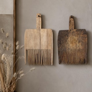 May include: Two antique wooden wool combs with long, thin, vertical teeth and a central handle. One comb is light-toned wood, while the other is a darker, weathered brown. Both feature a small hole at the top of the handle for wall mounting.