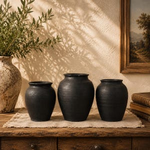 Può includere: Tre vasi in ceramica nera di diverse dimensioni sono esposti su una superficie di legno. I vasi hanno una finitura opaca e strutturata e una forma semplice e arrotondata. Lo sfondo comprende una parete dai toni neutri e un dipinto incorniciato.