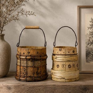 Puede incluir: Dos cubos cilíndricos de madera con asas de metal y motivos florales decorativos. El cubo izquierdo tiene un acabado marrón oscuro con una rejilla de alambre, mientras que el derecho es de color más claro con dos bandas horizontales de madera.
