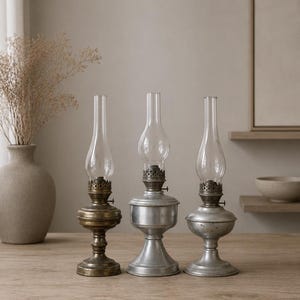 May include: Three oil lamps with clear glass chimneys and metal bases. The left lamp has a dark brass finish, while the center and right lamps feature a silver-toned metallic finish. Each lamp has a burner mechanism and a flared pedestal base.