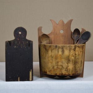 May include: A rustic kitchen set featuring a dark, rectangular cutting board with a handle and a wooden utensil holder. The holder contains wooden spoons and has a decorative top. The items are made of wood and have a vintage aesthetic.