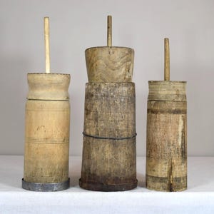 May include: Three antique wooden butter churns of varying sizes. The churns are cylindrical with a wooden lid and a long wooden handle. The wood has a natural, aged appearance, with visible grain and some wear. The churns are displayed on a white surface.