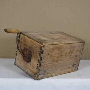 Puede incluir: Una caja de madera con una manivela. La caja es de madera marrón claro y tiene un aspecto rústico. La manivela es de metal y tiene un mango de madera.
