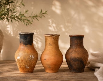 Puede incluir: Tres jarrones de terracota hechos a mano, con una paleta de colores cálidos y terrosos. Los jarrones tienen una forma clásica y redondeada con bordes ligeramente ensanchados. Las texturas y los tonos varían, con un borde oscuro en un jarrón.