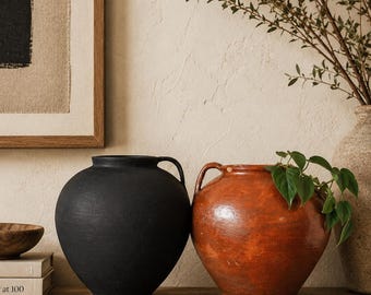 May include: Two ceramic vases sit on a wooden shelf. One is matte black, the other is a reddish-brown with a handle. Green plants are in the vases and a framed artwork is in the background.