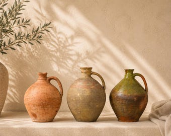 Könnte beinhalten: Drei irdene Krüge mit Henkeln in Orange-, Beige- und Grüntönen stehen auf einer neutralen Oberfläche. Links steht eine Vase mit Olivenzweigen. Der Hintergrund ist eine strukturierte, beige Wand mit Schatten.