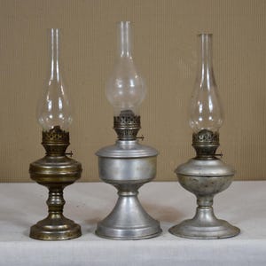 May include: Three vintage oil lamps with glass chimneys and metal bases. The lamps have different designs, with one featuring a brass base and the others silver-toned. They are displayed on a white surface against a neutral background.