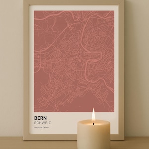 Puede incluir: Una impresión enmarcada del mapa de Berna, Suiza, en color rojo apagado. El mapa detalla las calles y vías fluviales. La impresión está etiquetada como "Bern Schweiz" y "Maptone Calma". Una vela encendida está frente a la impresión.