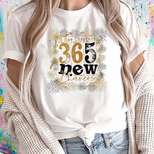 May include: White t-shirt with the text "365 new Chances" in a gold and black design. The design includes the text "12 New Chapters" and a decorative border of gold and silver starbursts.