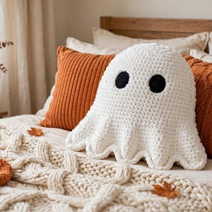 May include: A white crochet ghost cushion with two round black eyes, positioned on a bed between two orange textured throw cushions. The ghost has a rounded top and a scalloped bottom edge, crafted from thick yarn with a visible knit texture.