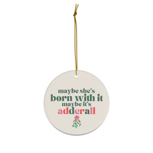 May include: A round white ceramic ornament with a green and pink design. The ornament features the text "maybe she's born with it maybe it's adderall" and a green mistletoe with a pink bow.