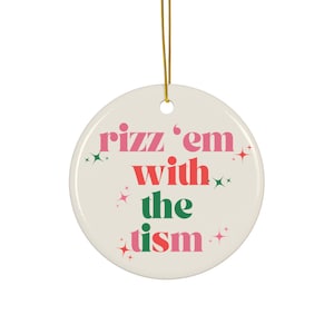 May include: A white ceramic Christmas ornament with the text "rizz 'em with the tism" in pink and green letters. The ornament has a gold string for hanging.