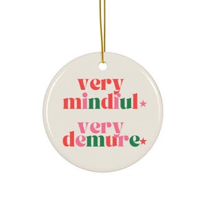May include: A round, off-white ceramic ornament with gold string. The ornament features the words "very mindful" and "very demure" in red, pink, and green text. A star is next to each word. The ornament is designed for hanging.