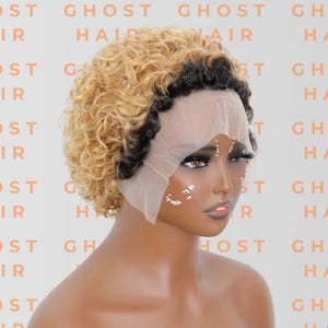 May include: A short, curly wig with a blonde and black color scheme. The wig is styled on a mannequin head with a lace front. The words "GHOST HAIR" are printed repeatedly in a peach color in the background.