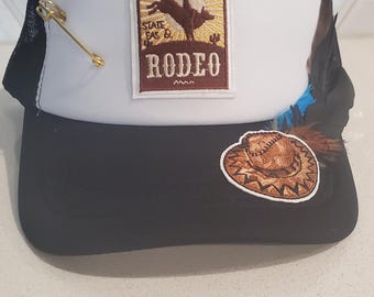 Może przedstawiać: Czarno-biała czapka typu trucker z naszywką "Cowboy State Fair & Rodeo". Czapka posiada złotą szpilkę w kształcie strzały, naszywkę z kapeluszem kowbojskim i ozdobne pióra. Ma czarny daszek i siatkowy tył.