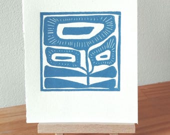 May include: A blue and white linocut print of an abstract floral design. The print is displayed on a small wooden easel. The artwork features a square composition with stylized flower shapes in a vibrant blue hue against a white background.
