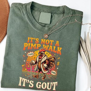 Op de afbeelding: Olijfgroen t-shirt met de tekst "IT'S NOT A PIMP WALK IT'S GOUT" en een cartoon haan met een jas en zonnebril. Een gouden ketting is over het shirt gedrapeerd. De haan houdt een wandelstok vast en heeft een dollarteken op het shirt.