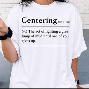 Puede incluir: Camiseta blanca con la palabra "Centering" en negro, seguida de su pronunciación fonética. Debajo, una definición dice: "(v.) El acto de luchar contra un montón de barro gris hasta que uno de vosotros se rinda."