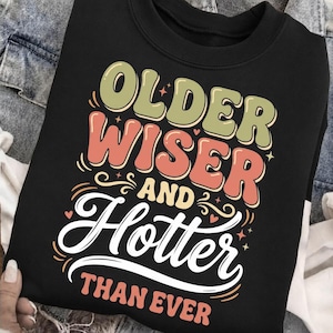 May include: Black sweatshirt with the text "OLDER WISER AND Hotter THAN EVER" in a retro font. The text is in shades of green, orange, and white. The sweatshirt is a casual style.