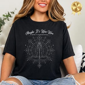 Pode incluir: Camiseta preta com o texto branco "Maybe It's Woo-Woo Maybe It's Quantum Physics". A camiseta apresenta um gráfico branco de uma pessoa meditando dentro de um design geométrico.