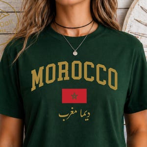 May include: A forest green short-sleeved t-shirt featuring the word MOROCCO in gold block letters, the red and green flag of Morocco, and the Arabic text ديما مغرب in gold. The shirt is paired with light blue distressed denim jeans.