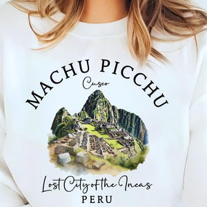 Może przedstawiać: Biała bluza z nadrukiem Machu Picchu, Zaginionego Miasta Inków, z napisem "Machu Picchu Cusco Peru". Projekt zawiera szczegółową ilustrację starożytnego miejsca. Bluza ma okrągły dekolt i długie rękawy.