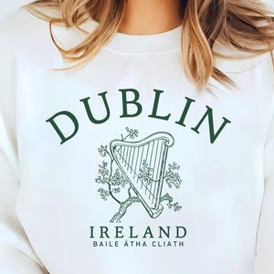 May include: White sweatshirt with the word "DUBLIN" in green arched text above a green harp illustration. Below the harp is the word "IRELAND" and "BAILE ÁTHA CLIATH" in green. The sweatshirt has a crew neck.