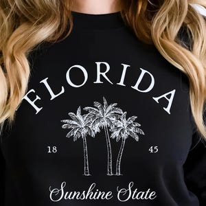 May include: Black sweatshirt featuring the word "FLORIDA" in white, arched across the top, with a graphic of three palm trees and the text "Sunshine State". The sweatshirt also has the numbers "18" and "45" printed below the palm trees.