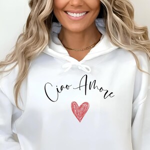 Peut inclure: Un sweat à capuche blanc avec un cordon de serrage et une poche avant. Le sweat à capuche porte l'inscription "Ciao Amore" en écriture noire au-dessus d'un cœur rouge. Le logo de la marque est dans le coin supérieur gauche.