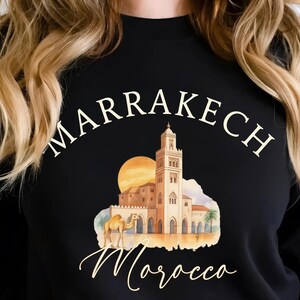 May include: Black sweatshirt featuring a watercolor illustration of a Moroccan scene with a mosque, camel, and sunset. The word "Marrakech" arches across the top, with "Morocco" in script below the image. The word "Black" is in the bottom right corner.