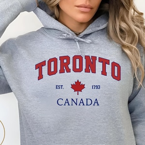 Könnte beinhalten: Sportgrauer Kapuzenpullover mit Vordertasche und Kapuze. Das Wort "TORONTO" ist in roten, gebogenen Buchstaben über "CANADA" und einem Ahornblatt aufgedruckt. Der Text "EST. 1793" ist ebenfalls auf dem Hoodie aufgedruckt.