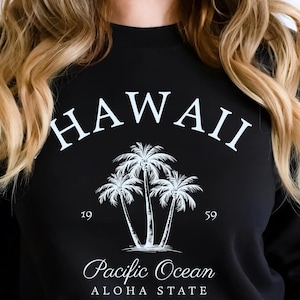 Peut inclure: Sweat-shirt noir avec un motif blanc. Le motif comprend le mot "HAWAII", un dessin de palmiers, les mots "Pacific Ocean" et "Aloha State", ainsi que les chiffres "19" et "59".