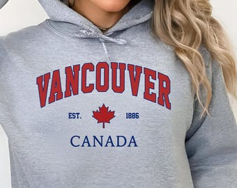 May include: A light grey hooded sweatshirt with a front pocket. The word "VANCOUVER" is arched across the chest in red letters, with "CANADA" below. A red maple leaf and the year "1886" are also featured. The sweatshirt is labeled "Sport grey".