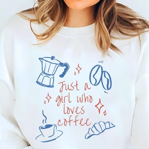 Peut inclure: Sweat-shirt blanc avec un motif sur le thème du café. Le design comprend une cafetière bleue, des grains de café, une tasse de café et un croissant, avec le texte "Just a girl who loves coffee" en rouge et bleu.