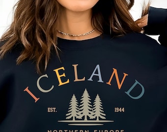 Puede incluir: Sudadera negra con la palabra "ICELAND" en letras arqueadas de colores, con un gráfico de tres pinos y el texto "EST. 1944 NORTE DE EUROPA". La sudadera tiene cuello redondo y mangas largas.