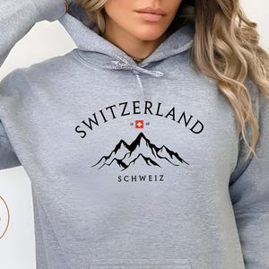 Peut inclure: Sweat à capuche gris sport avec une poche avant et une capuche à cordon. Le devant porte l'inscription "SWITZERLAND" au-dessus d'un motif de montagne et le mot "SCHWEIZ". Le drapeau suisse est intégré au design. Le logo Atlas Doll Crafts est en bas à gauche.