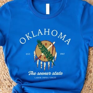 Peut inclure: Un t-shirt bleu roi avec le mot "OKLAHOMA" en texte blanc arqué. En dessous, un emblème circulaire avec un motif amérindien. Le t-shirt comprend également le texte "The sooner state" et "Labor omnia vincit".