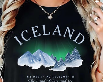 Puede incluir: Camiseta negra con la palabra "ICELAND" en blanco arqueada en la parte superior. Debajo, un gráfico que representa montañas, árboles y una aurora verde y blanca. También está impreso el texto "64.9631° N, 19.0208° W" y "The Land of Fire and Ice".