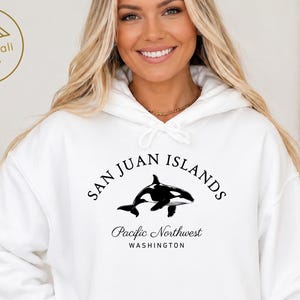 Peut inclure: Un sweat-shirt à capuche blanc avec un motif noir représentant une orque au centre de la poitrine. Le texte au-dessus de l'orque indique SAN JUAN ISLANDS, tandis que le texte en dessous indique Pacific Northwest WASHINGTON en polices variées.