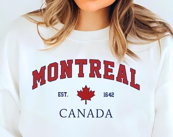 May include: White sweatshirt with the word "MONTREAL" in red arched lettering, "EST. 1642" and a red maple leaf graphic. Below, the word "CANADA" is in blue. The sweatshirt is a crew neck style.