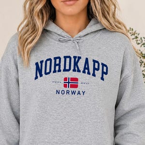 Peut inclure: Un sweat à capuche gris sport arborant le texte NORDKAPP en lettres arquées bleu foncé au-dessus d'une petite icône de drapeau norvégien et du mot NORWAY. Le sweat comprend une poche kangourou et des cordons de serrage gris assortis.
