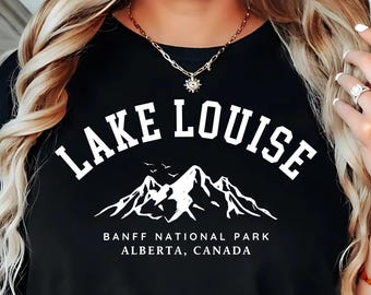 May include: Black t-shirt featuring the white text "Lake Louise" above a mountain graphic and the text "Banff National Park Alberta, Canada." The shirt has a crew neck.