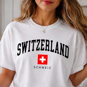 May include: A white short-sleeved t-shirt featuring the word SWITZERLAND in black arched lettering above a red square containing a white cross, with the word SCHWEIZ printed in black underneath the square.