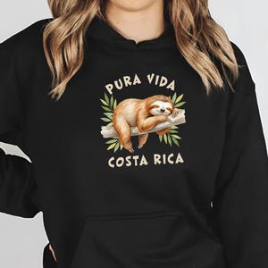 Peut inclure: Sweat-shirt à capuche noir avec une illustration de paresseux sur une branche entourée de feuilles vertes. Le texte "Pura Vida Costa Rica" est au-dessus et en dessous du paresseux. Le sweat-shirt a une poche avant et une capuche à cordon.