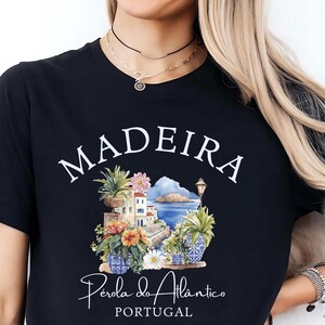 Puede incluir: Camiseta negra con un diseño gráfico colorido que presenta la palabra "MADEIRA" en blanco, junto con una ilustración de una escena costera. También se incluye el texto "Pérola do Atlântico PORTUGAL".